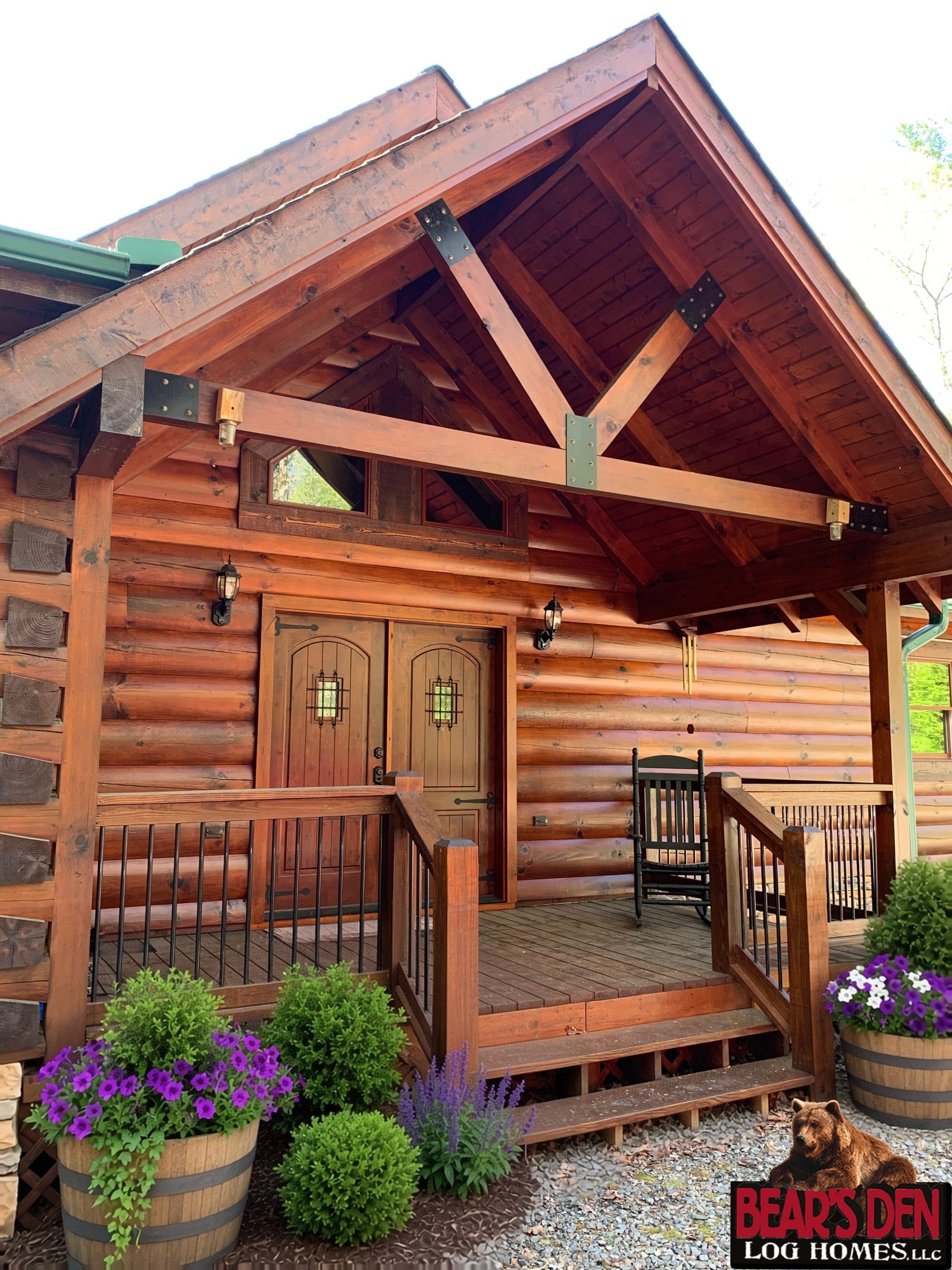 Timber framed entry porch with double wood doors on a log cabin, with barrel planters of purple petunias by the steps